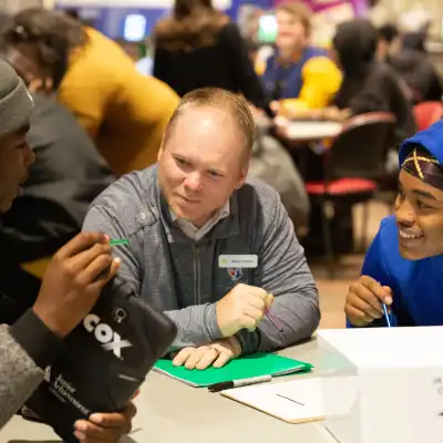 Two high school students and an adult volunteer sitting at a table. One student is showing the volunteer a tablet with the JA Finance Park simulation